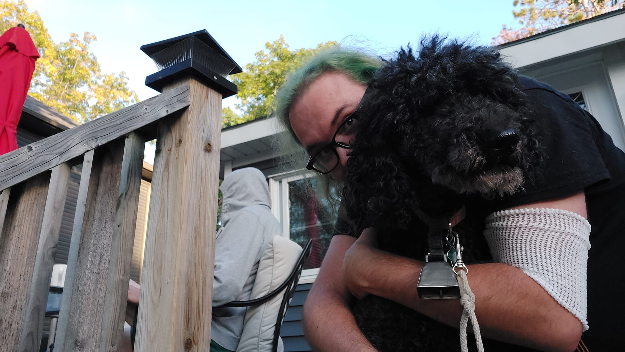 A man with long green gives a black standard poodle a hug, holding her in place to take the photo